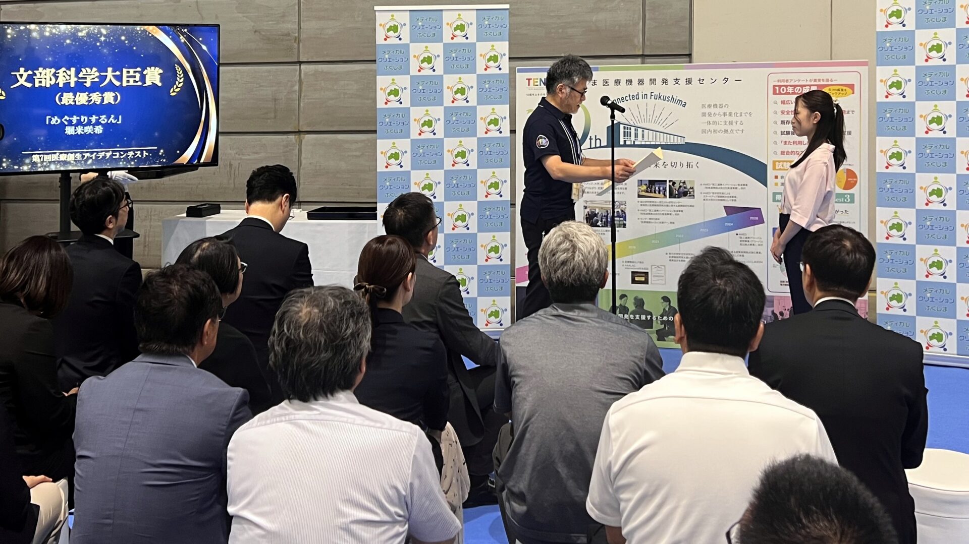 文部科学大臣賞の表彰式。右が堀米さん。左の画面に賞名と受賞作「めぐすりするん」の名前が映る